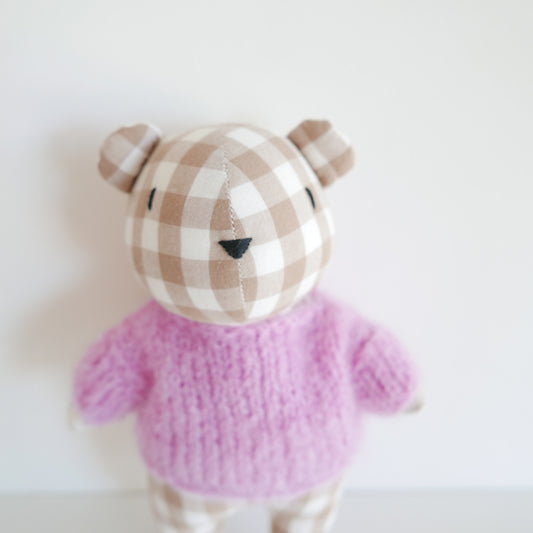 Small teddy bear with a checkered hat and pink sweater on a light background