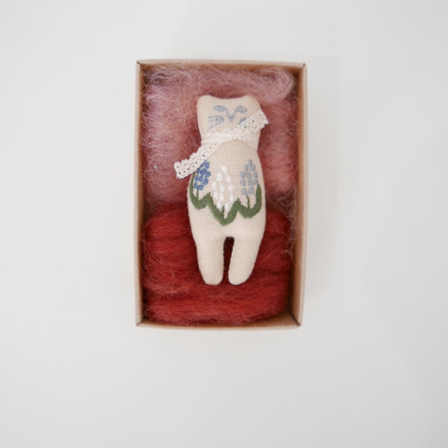 Small embroidered doll in a box with red fiber on a light gray background