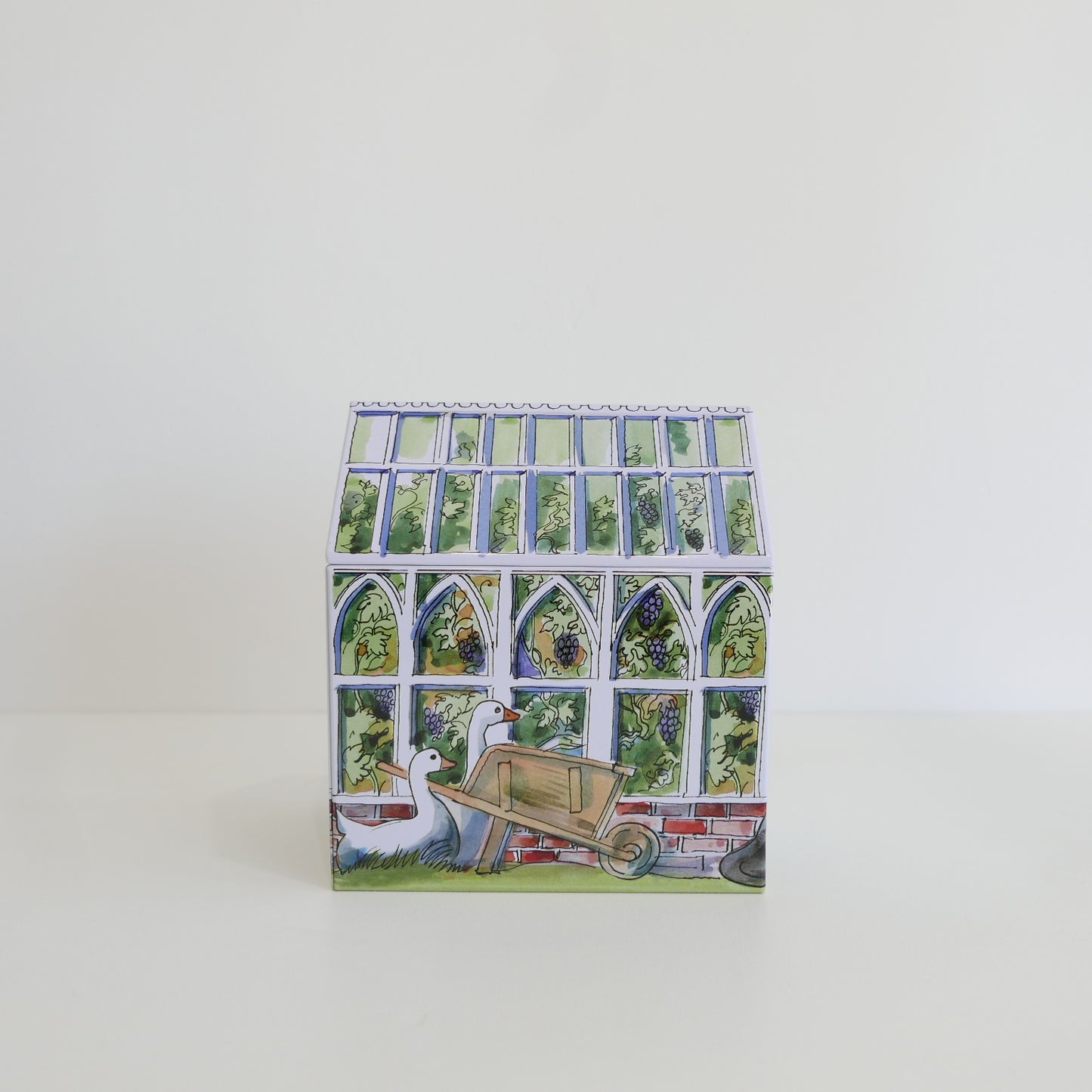 Small tin box with a decorative design of a garden scene, including a duck and a wheelbarrow, on a white background.