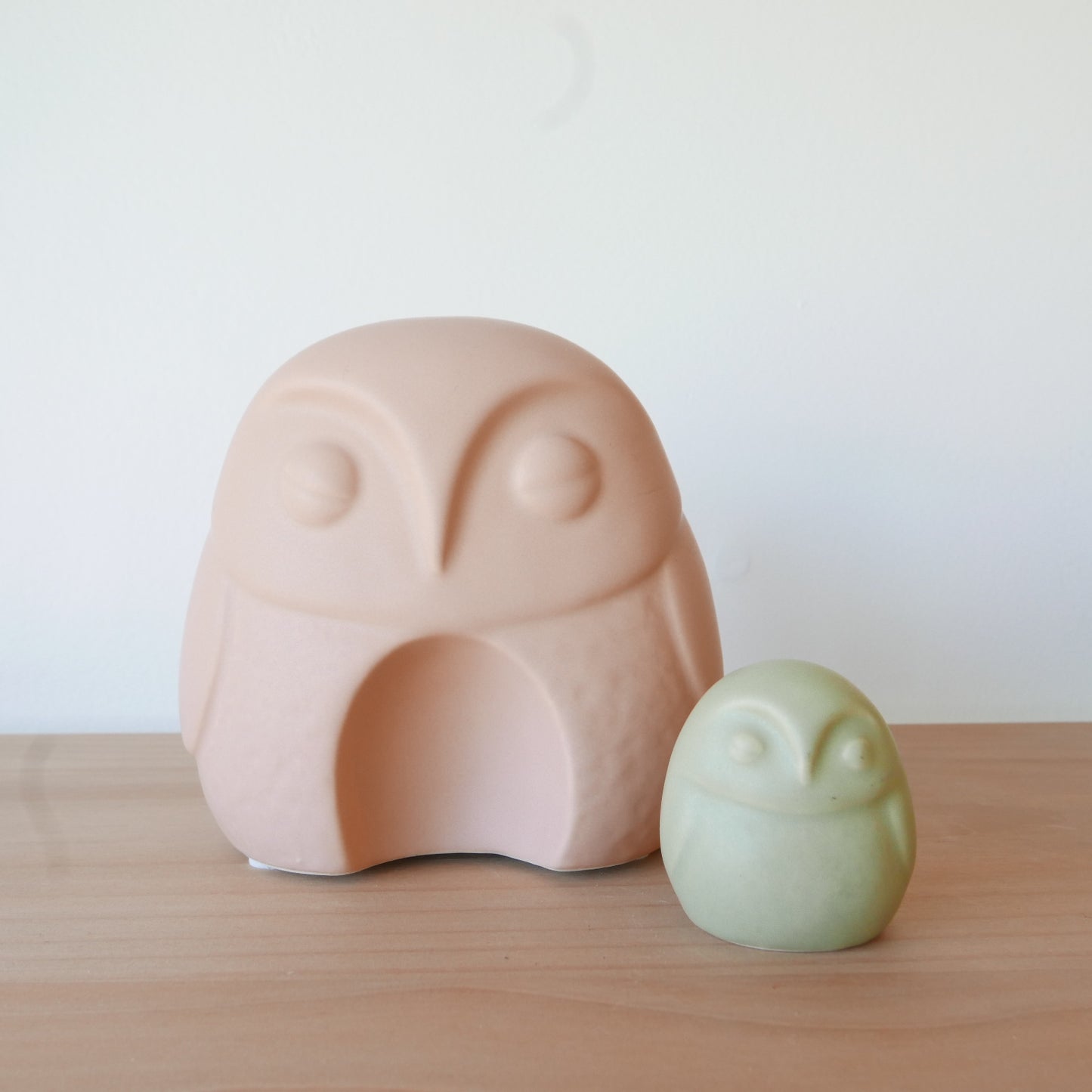 Two owl-shaped objects, one pink and one green, on a wooden surface with a white background.