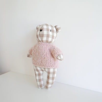 Stuffed teddy bear toy wearing a pink sweater and checkered pants on a white background