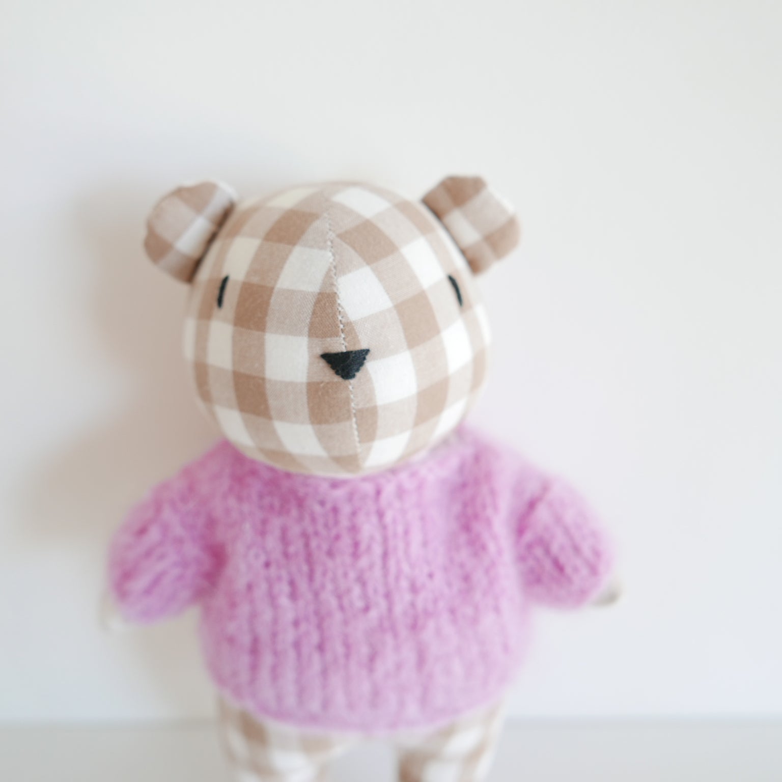 Small teddy bear with a checkered hat and pink sweater on a light background