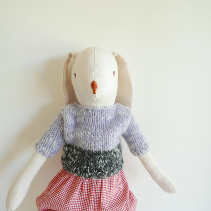 Stuffed rabbit toy wearing a knitted sweater and checkered skirt on a white background