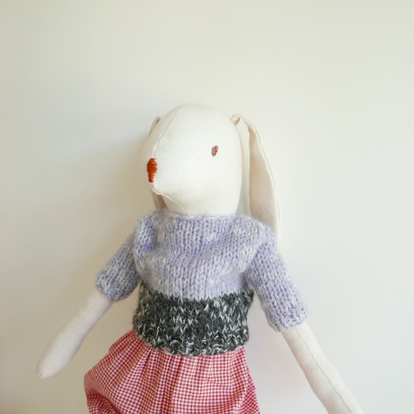 White rabbit plush toy wearing a knitted sweater and red checkered skirt on a light background
