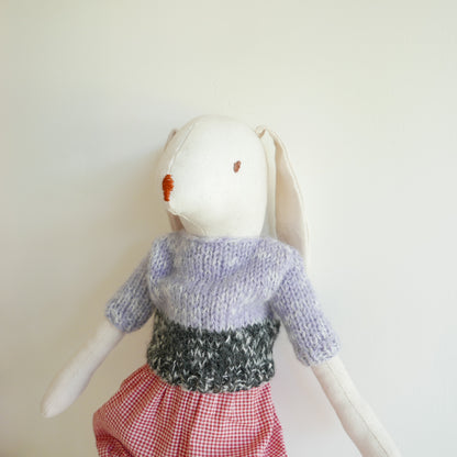 White rabbit plush toy wearing a knitted sweater and red checkered skirt on a light background